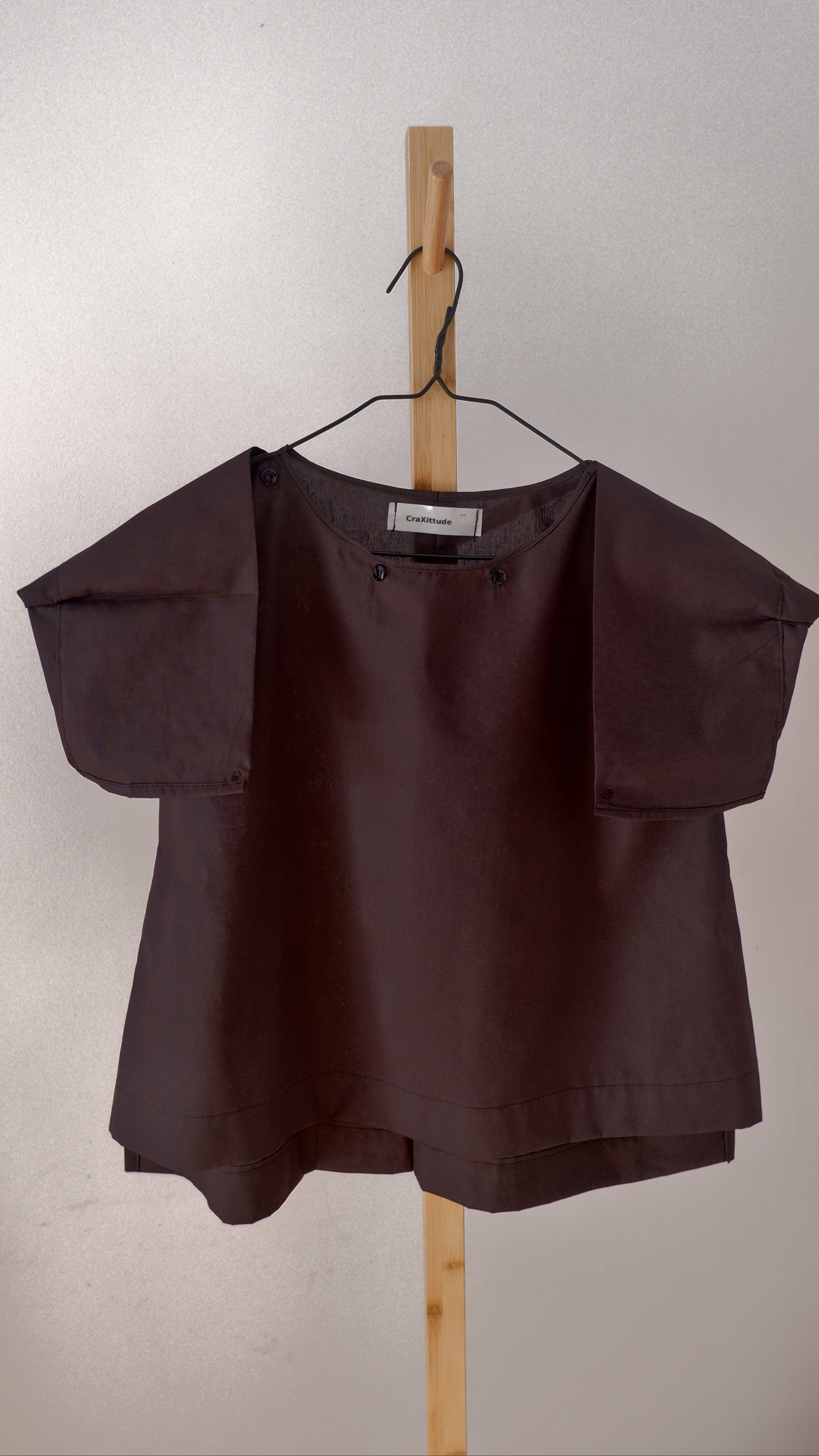 A black short-sleeve blouse with structured shoulder details, hanging on a wooden hanger from a wooden stand against a plain off-white wall