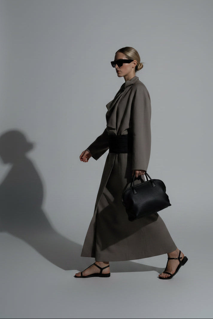 A woman wearing a flowing taupe-gray draped trench coat with a wide black belt, black sunglasses, and carrying a black shoulder bag.